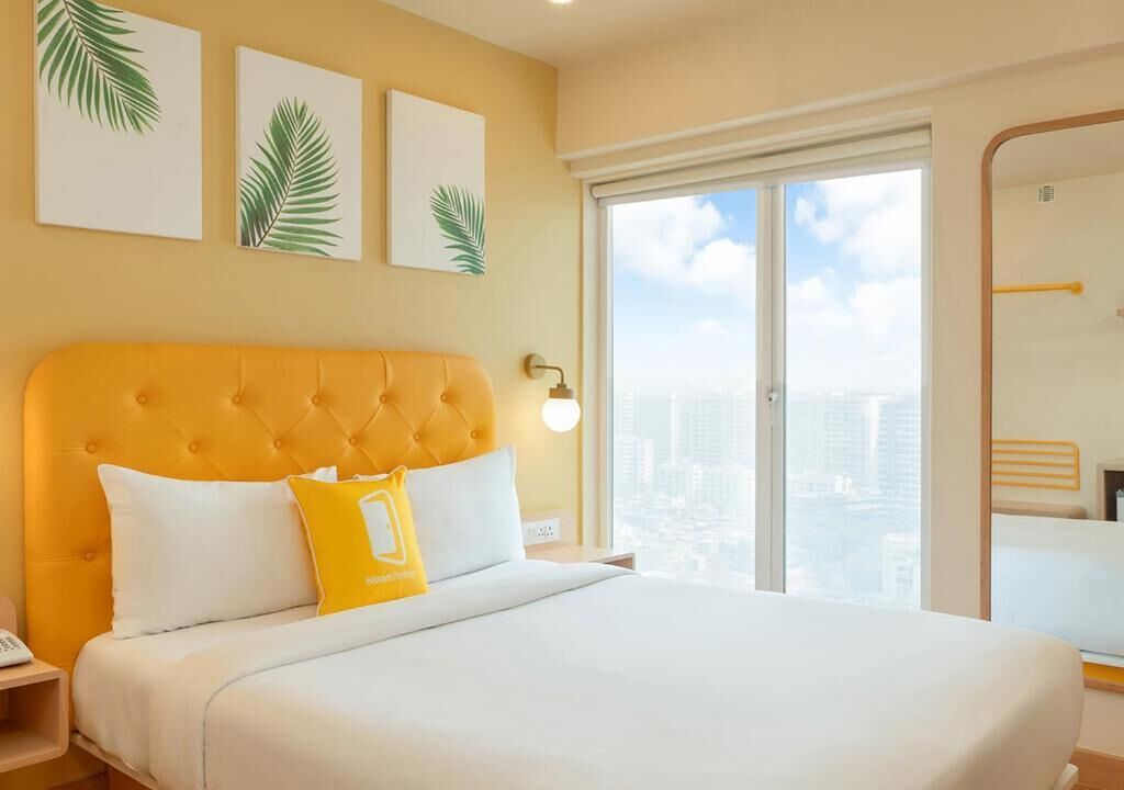 A hotel room with a large bed, a city view, and artwork on the wall.