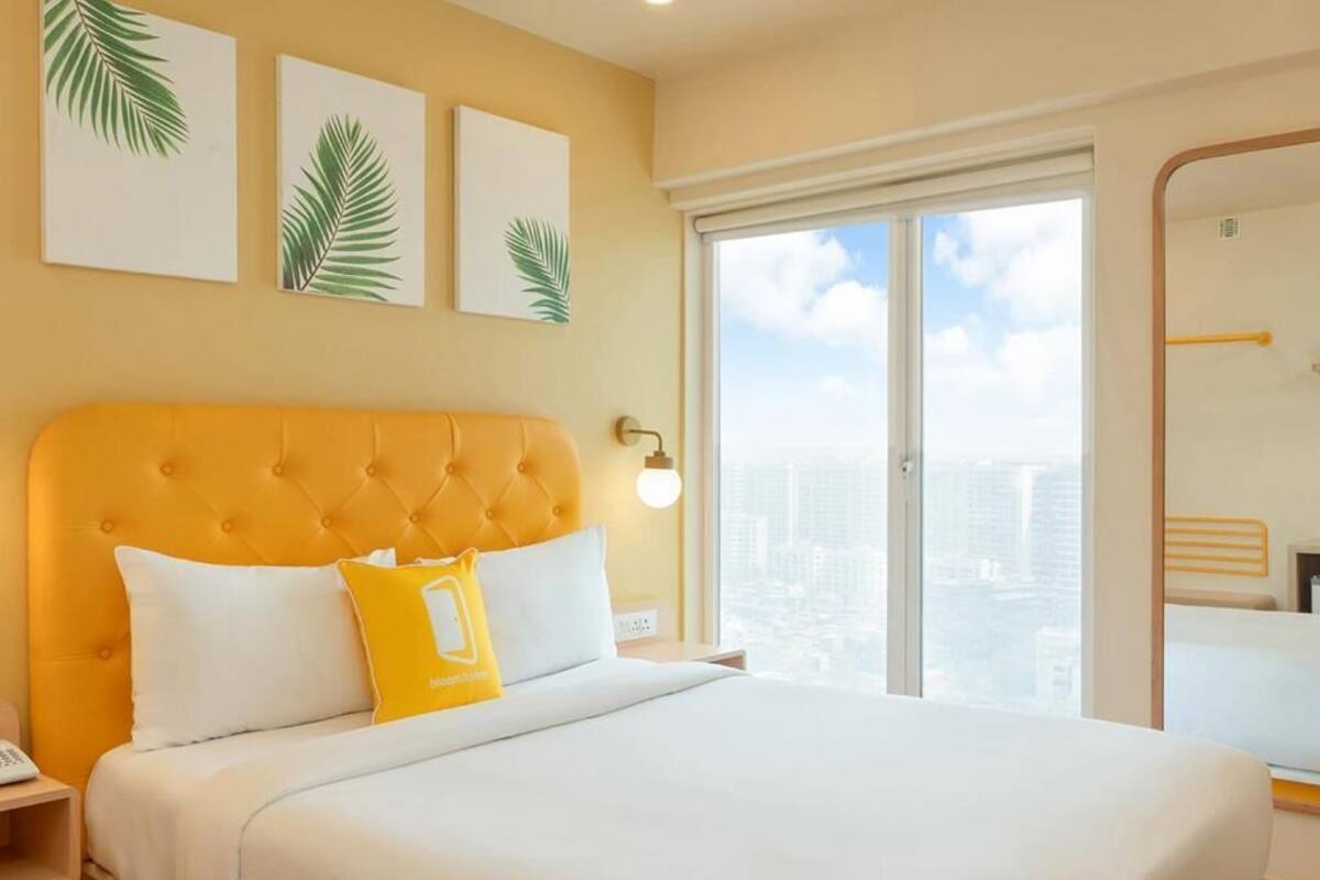 A hotel room with a large bed, a city view, and artwork on the wall.