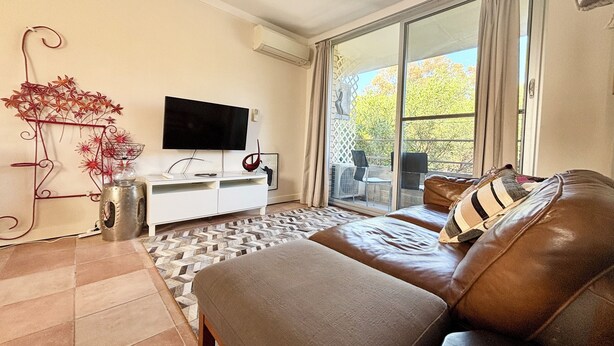 A modern living room with a large flat-screen TV, a white entertainment unit, a brown leather sofa, and a view of trees through a sliding glass door. - City Escape Getaway Subiaco 1Bedrm Unit (Subiaco)