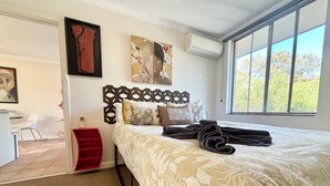 A bedroom with a large window, a bed with a patterned bedspread, a red chair, and a desk with a chair. - City Escape Getaway Subiaco 1Bedrm Unit (Subiaco)