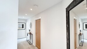Een gang met een houten deur, een lijst met een afbeelding aan de muur, en een tafeltje met een vaas.