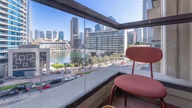 A balcony with a red chair, overlooking a marina with boats, a cityscape, and a road with vehicles. - New Listing | JBR Beach Gem | Czechin | 1BD (Dubai)