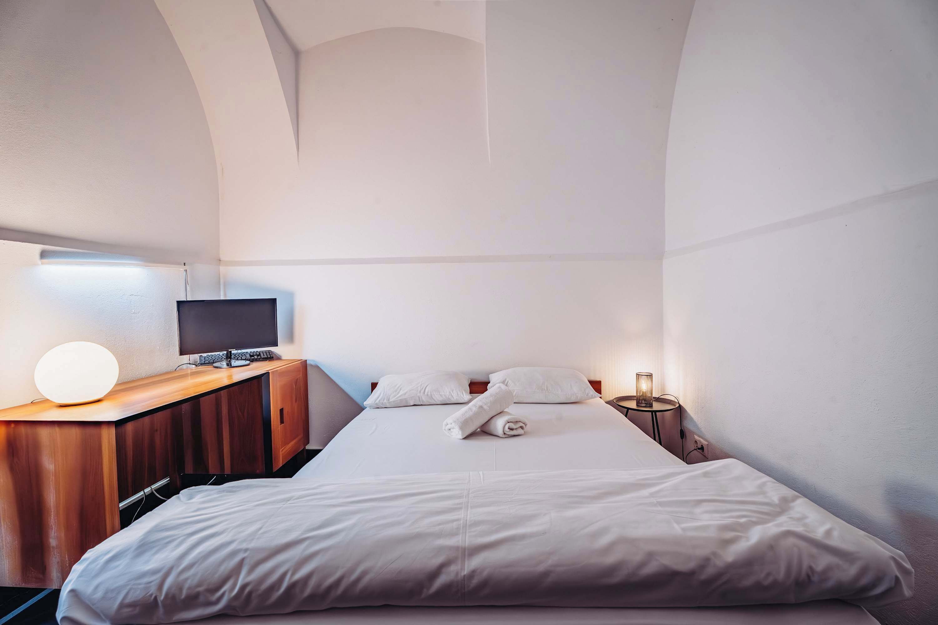 Ein Hotelzimmer mit einem Bett, einem Schreibtisch mit Computer, einer Lampe und einem Nachttisch ebenfalls mit Lampe.