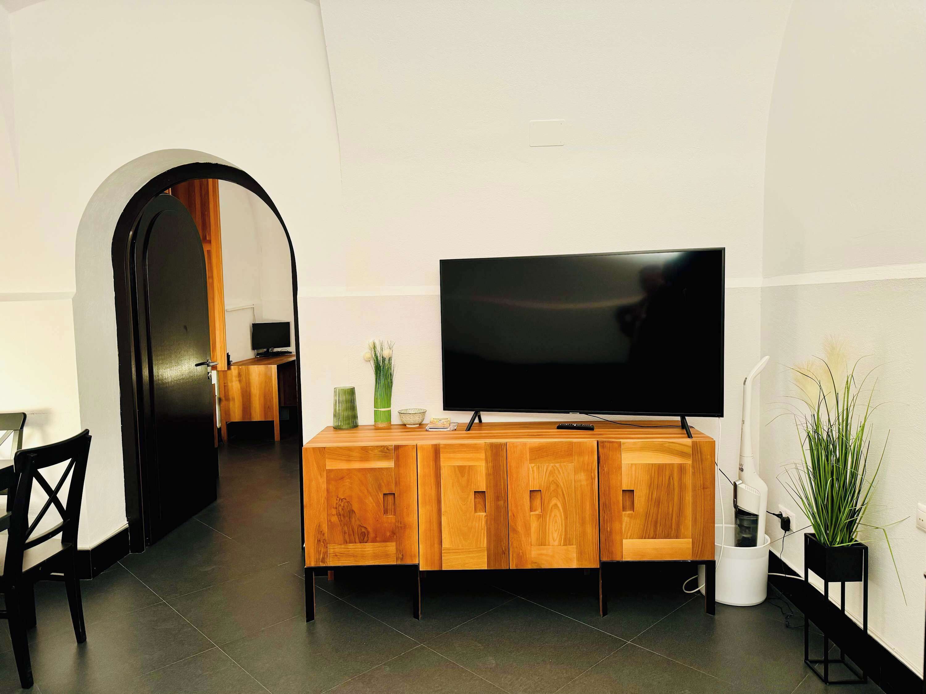A modern living room with a flat-screen TV mounted on the wall, a wooden entertainment unit, and a dining area with a chair and table.