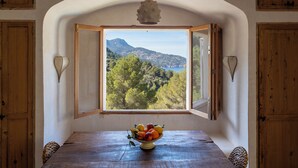 Una mesa de comedor con un frutero, vista a un paisaje montañoso a través de una ventana abierta, y paredes de madera.