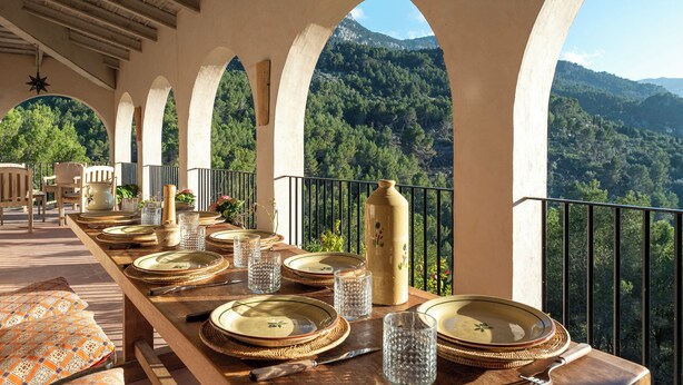 Una mesa de comedor dispuesta con platos, vasos y cubiertos, con vistas a un paisaje montañoso.