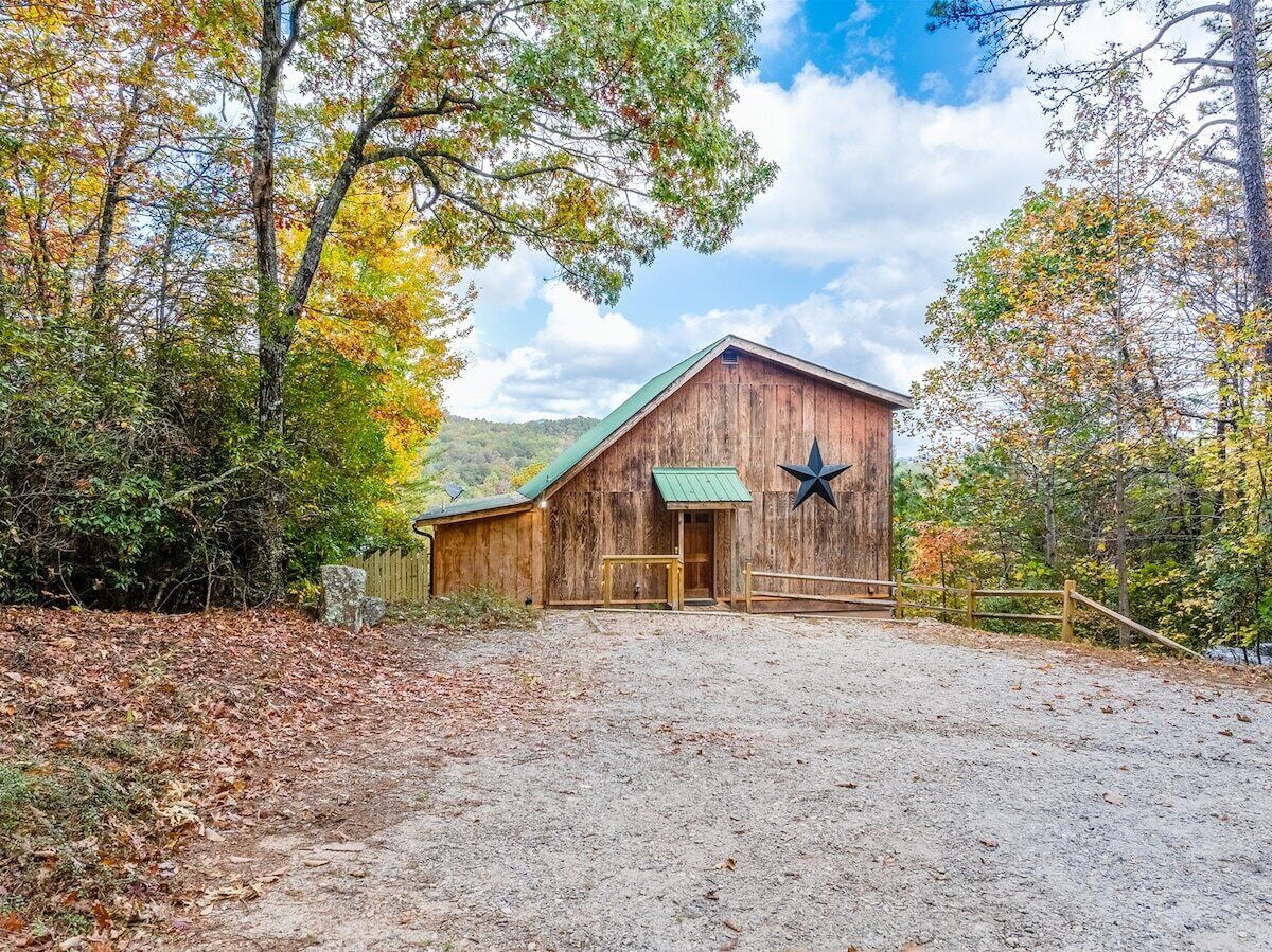 A rustic barn with a star decoration, surrounded by trees and a gravel driveway.