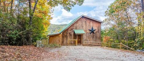 A rustic barn with a star decoration, surrounded by trees and a gravel driveway.