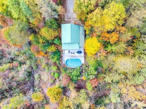 An aerial view of a house with a swimming pool, surrounded by dense trees with autumn foliage.