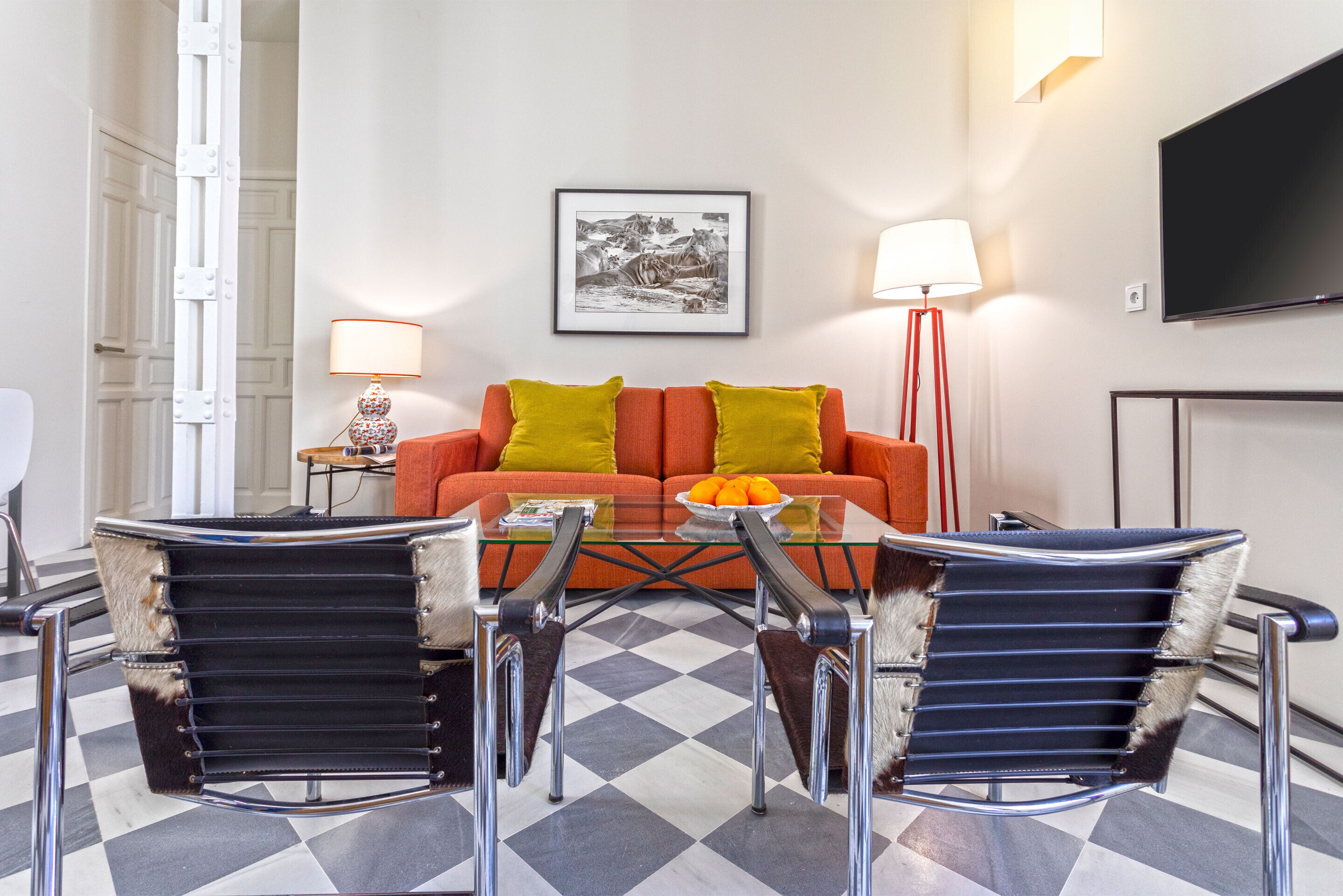 A modern living room with a glass coffee table, two leather chairs, a sofa with orange cushions, a framed picture on the wall, and a flat-screen TV.