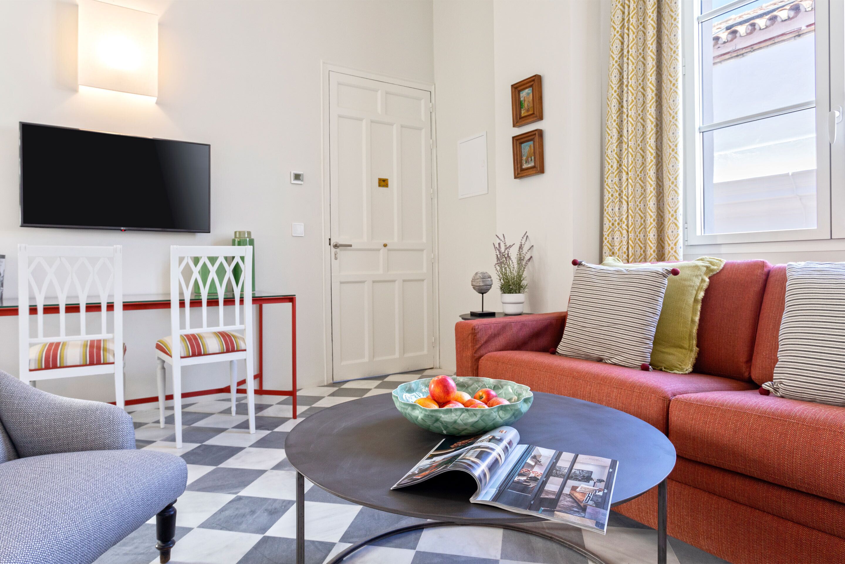 Una sala de estar con piso a cuadros, un sofá, una mesa de centro con una revista, un televisor en la pared y una puerta.