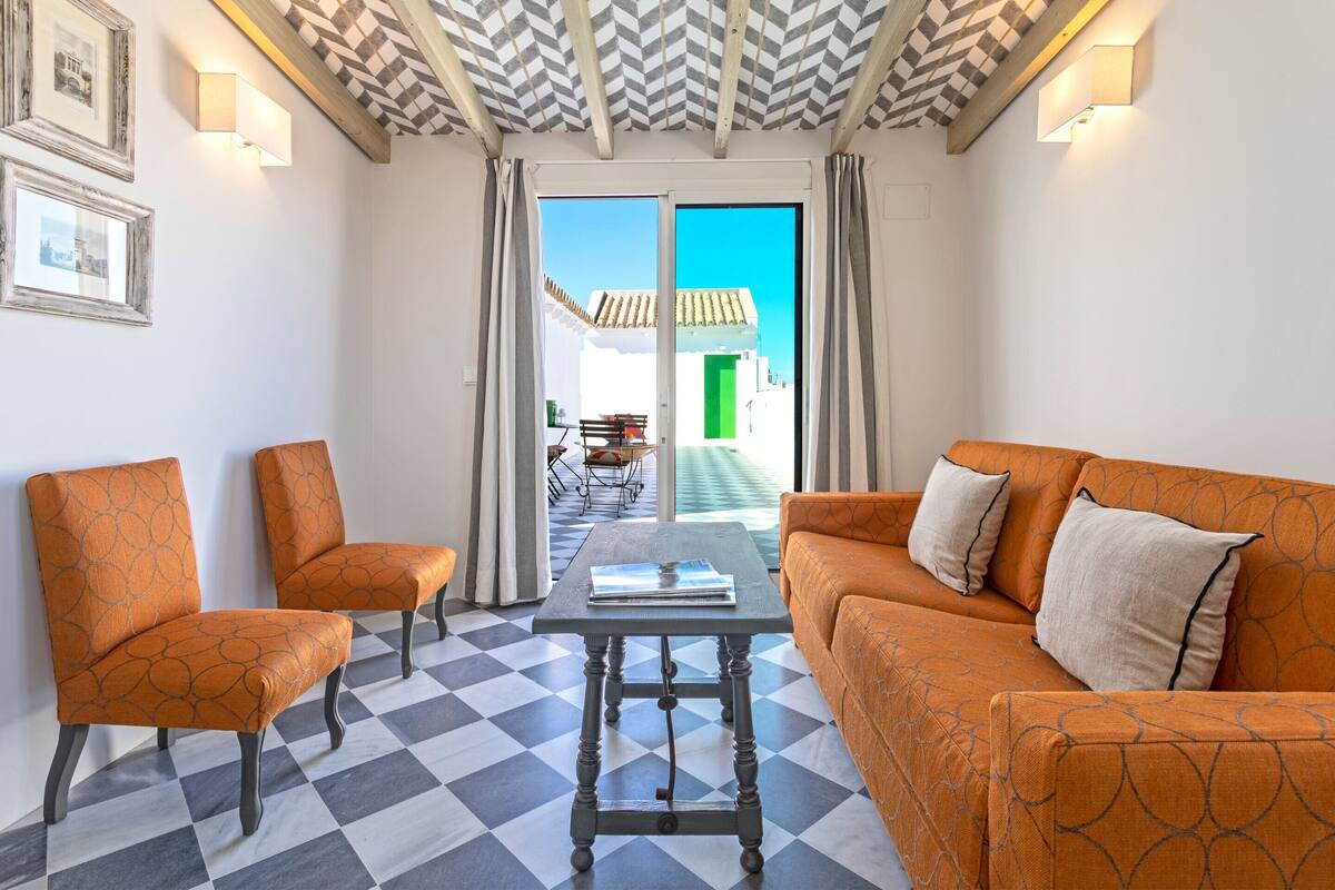 Una habitación con suelo a cuadros, dos sillones naranjas, un sofá y una mesa con un libro.