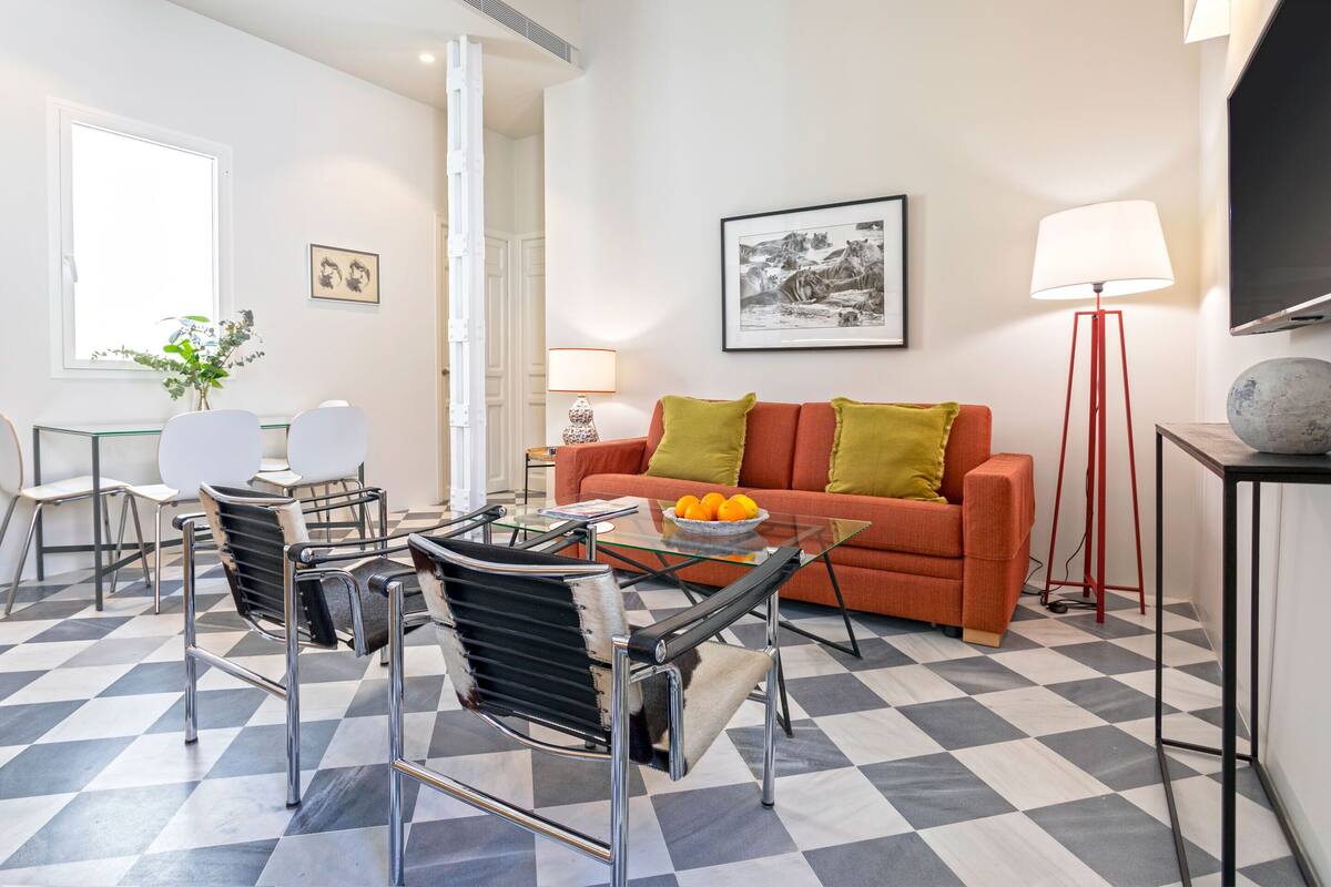 Una sala de estar moderna con suelo a cuadros, una mesa de centro de vidrio, un sofá con cojines, una mesita auxiliar, una lámpara y un cuadro en la pared.