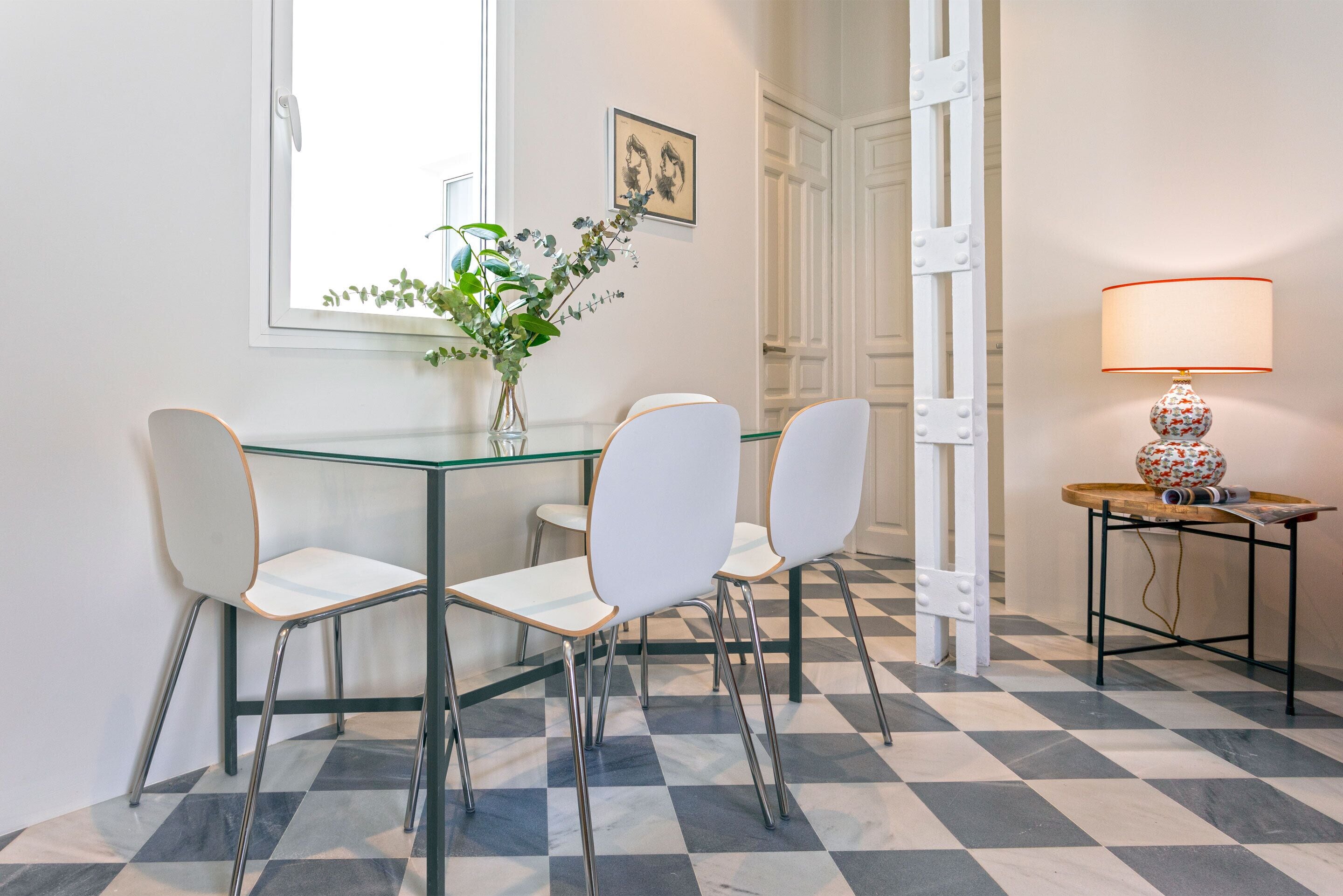 A dining area with a glass table, white chairs, a vase with greenery, a lamp, and a checkered floor.