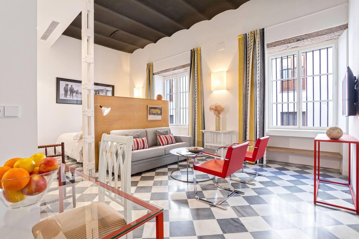 Una sala de estar moderna con piso a cuadros, un sofá, una butaca roja, una mesa de centro de vidrio y un cuenco con fruta.