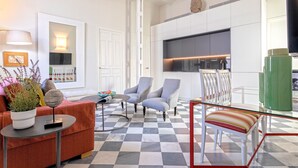 Ein modernes Wohnzimmer mit Schachbrettmuster-Boden, einer Couch, zwei Sesseln, einem Glastisch sowie einer Küchenzeile mit Schränken und einem Fernseher.