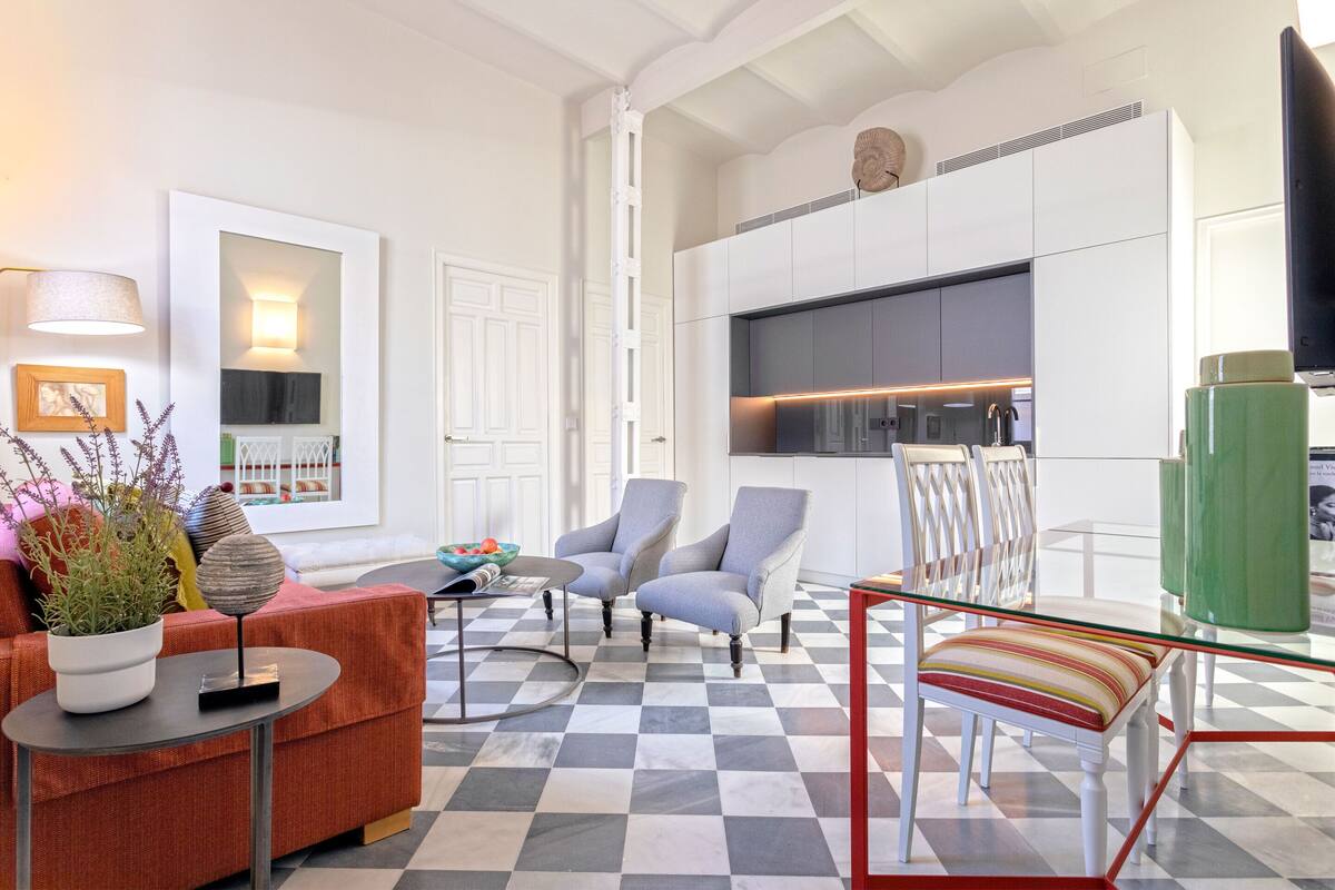 Una sala moderna con piso a cuadros, un sofá, dos sillones, una mesa de vidrio y una cocina con gabinetes y un televisor.