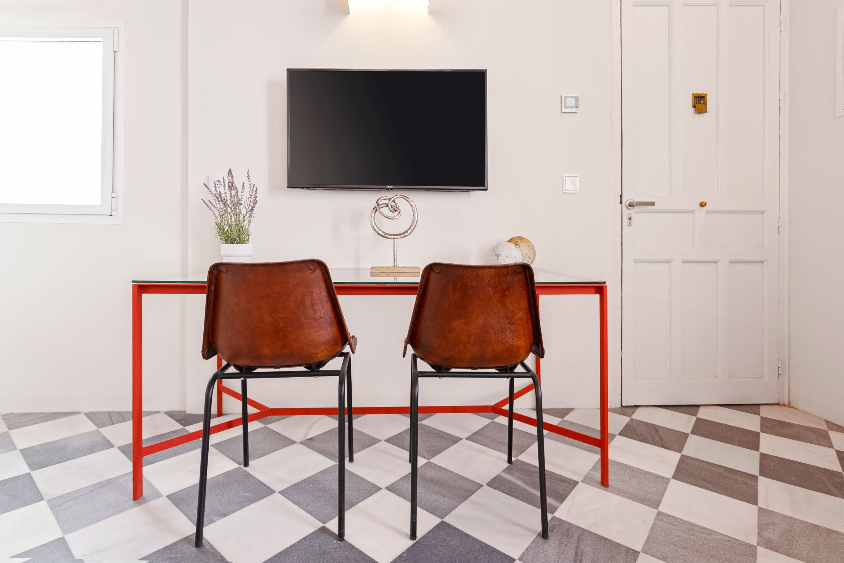 A room with a checkered floor, a red-framed desk, two brown chairs, a flat-screen TV mounted on the wall, and a white door.