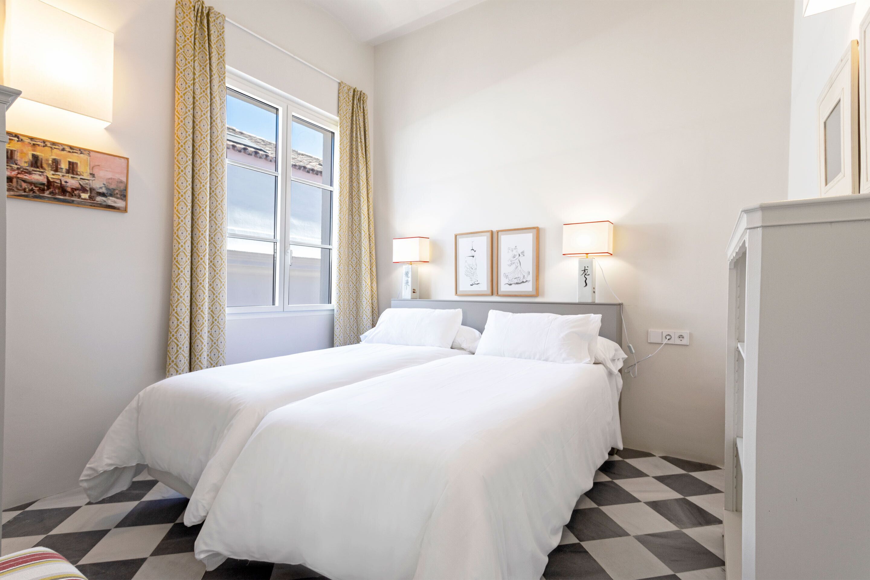 A hotel room with two beds, a checkered floor, a window with curtains, wall-mounted lamps, and a refrigerator.