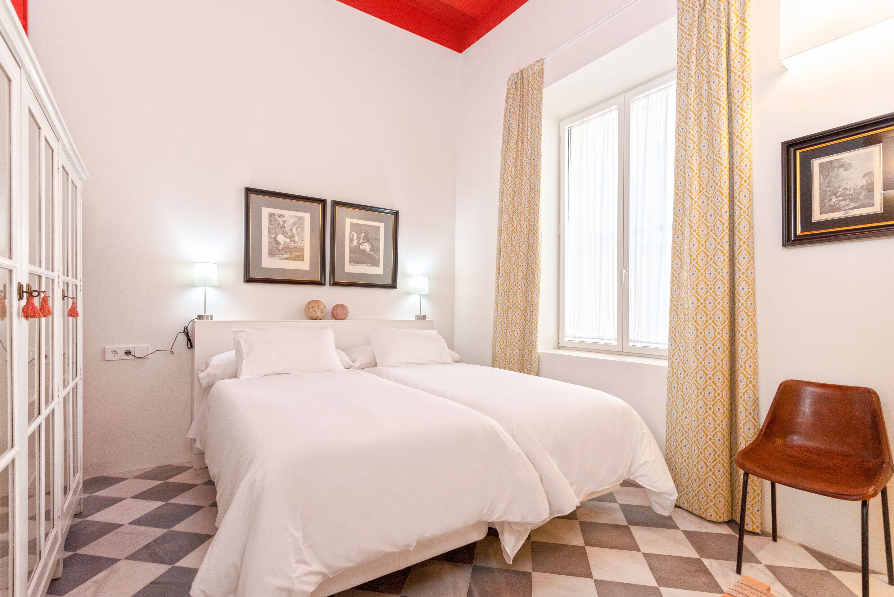 A bedroom with two beds, a window with patterned curtains, and a chair.