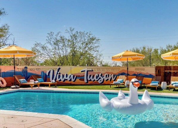 Ein Schwimmbecken mit Liegestühlen und Sonnenschirmen, ein Wandgemälde mit dem Schriftzug "Viva Tucson" und ein Schwaneinhorn-Luftmatratze.