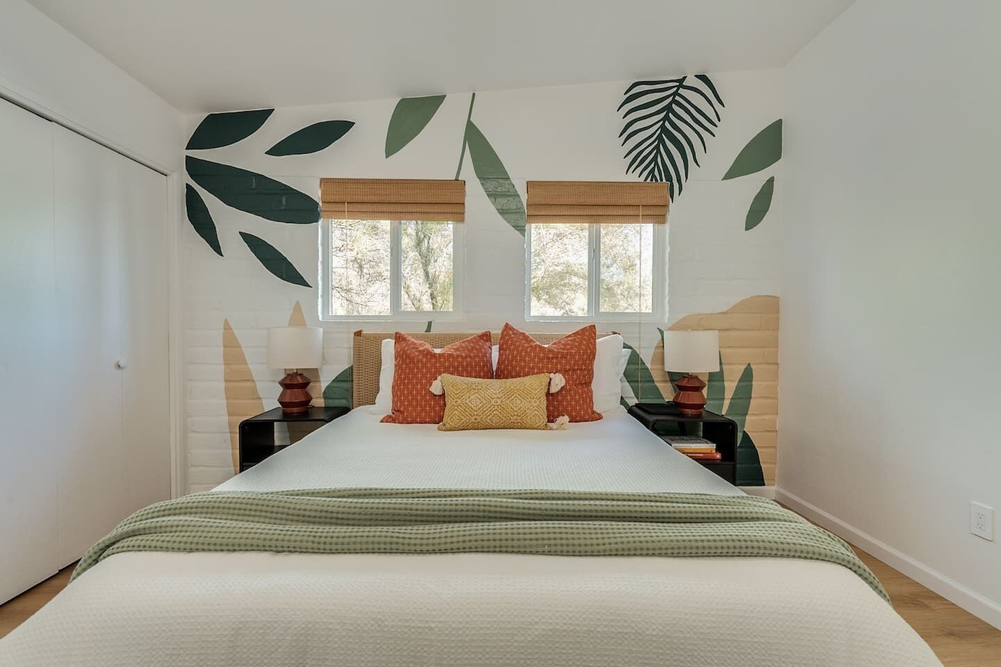 Un dormitorio con cama, dos mesitas de noche, ventana con vistas y decoración de pared con estampado de hojas.
