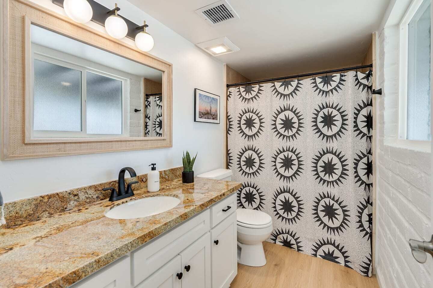 A bathroom with a granite countertop, a white sink, a black faucet, a white toilet, and a shower with a sunburst pattern wall.