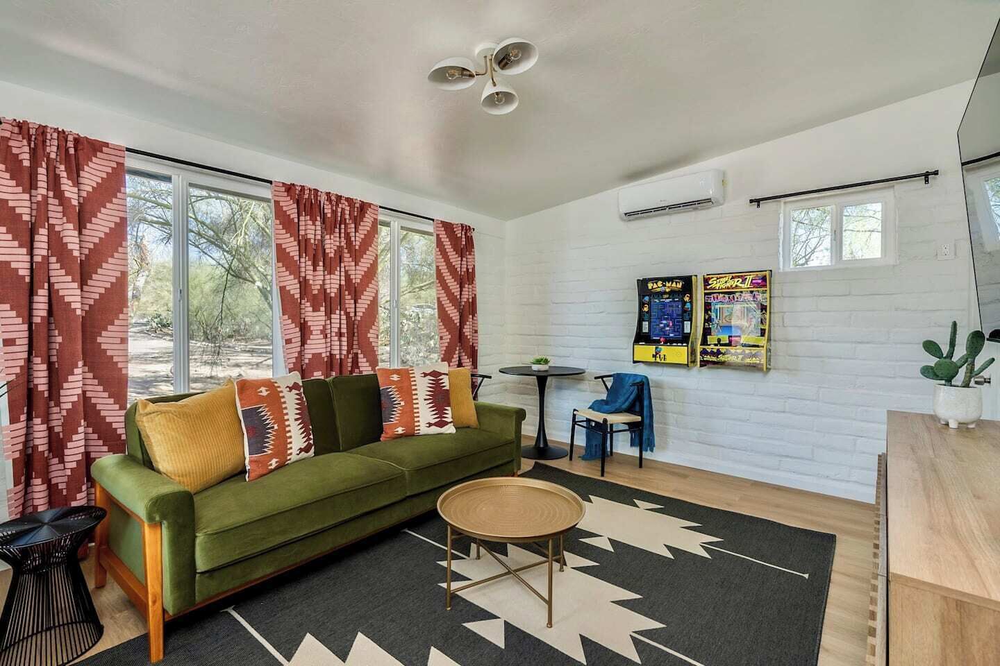 A living room with a green sofa, patterned cushions, a round coffee table, and a black and white rug. There are two windows with red curtains, an air conditioning unit, and a small table with a chair.