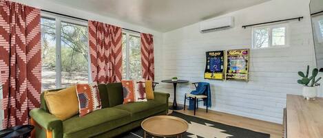 A living room with a green sofa, patterned cushions, a round coffee table, and a black and white rug. There are two windows with red curtains, an air conditioning unit, and a small table with a chair.