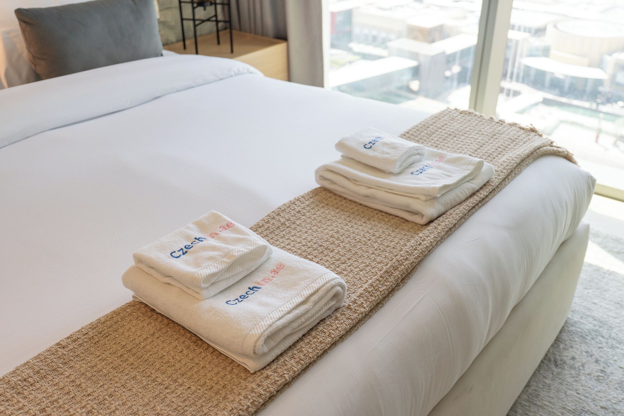 A neatly made bed with white linens and two folded towels with "Czech" embroidered on them, placed on a textured beige rug.