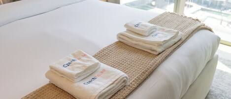 A neatly made bed with white linens and two folded towels with "Czech" embroidered on them, placed on a textured beige rug.