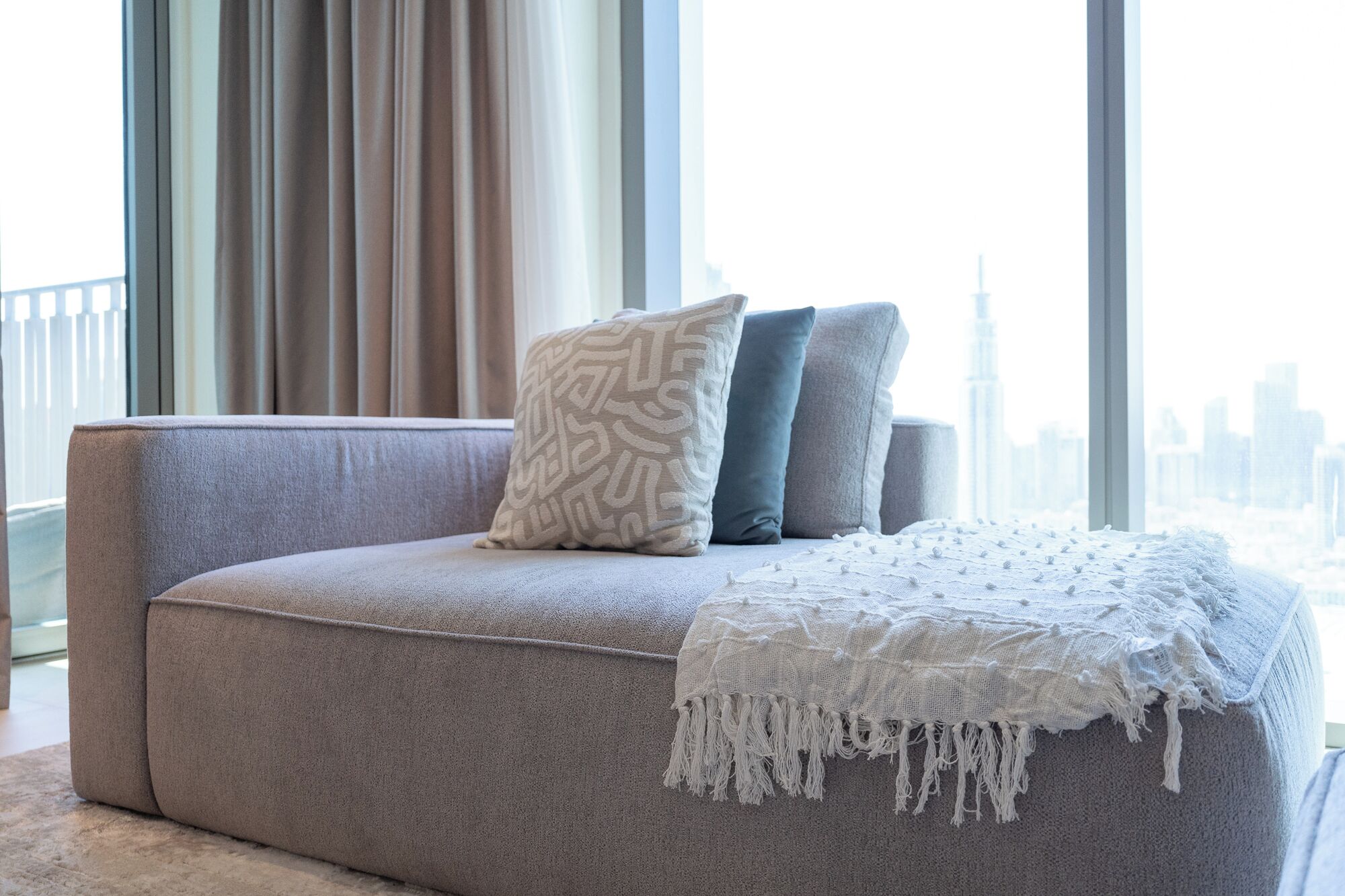 A modern sofa with patterned and solid pillows, a white fringed throw, and a cityscape view through large windows.