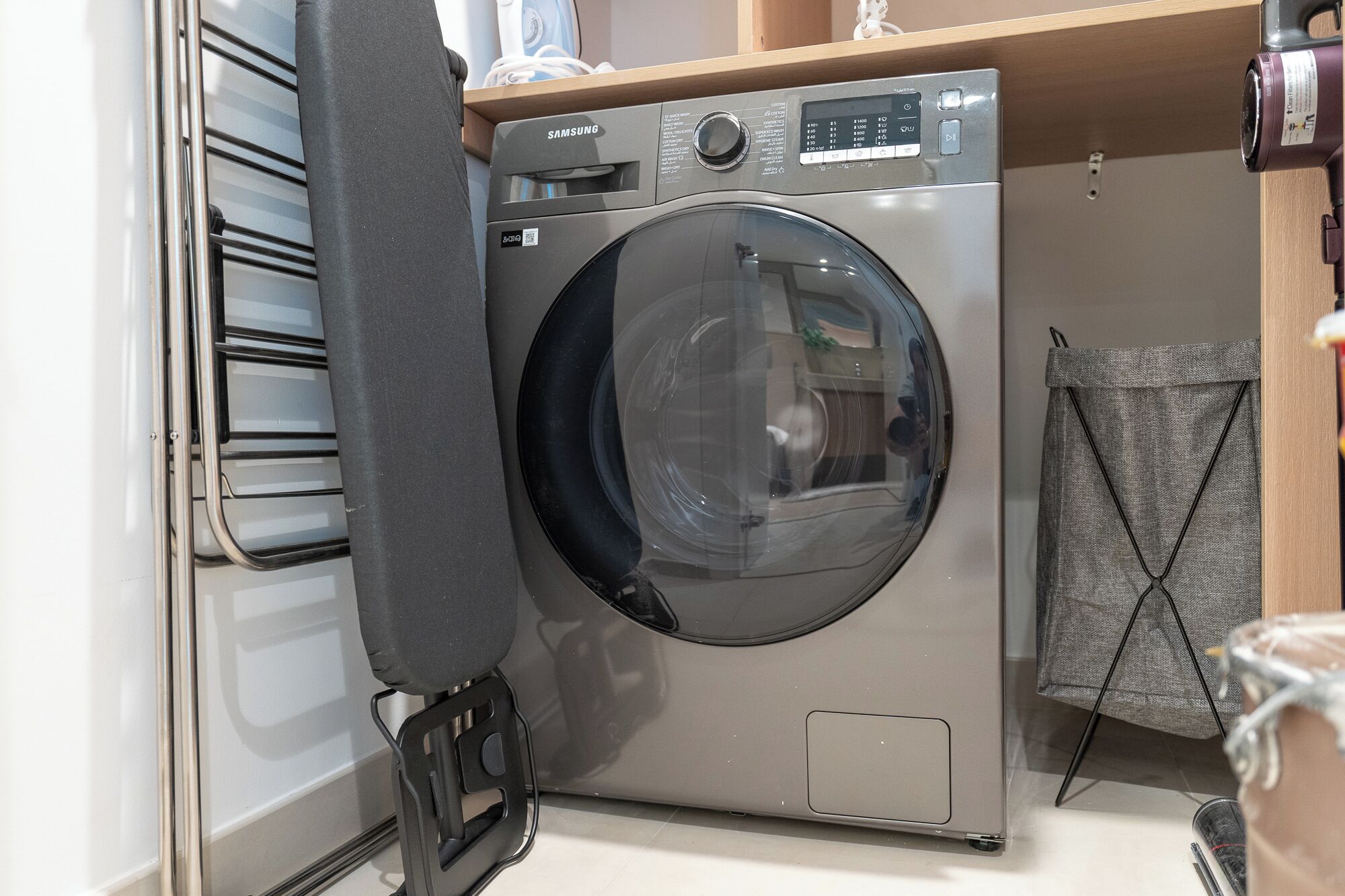 A compact, stainless steel front-loading washing machine with a control panel, located in a small laundry area with a wooden shelf above and a towel rack to the right.