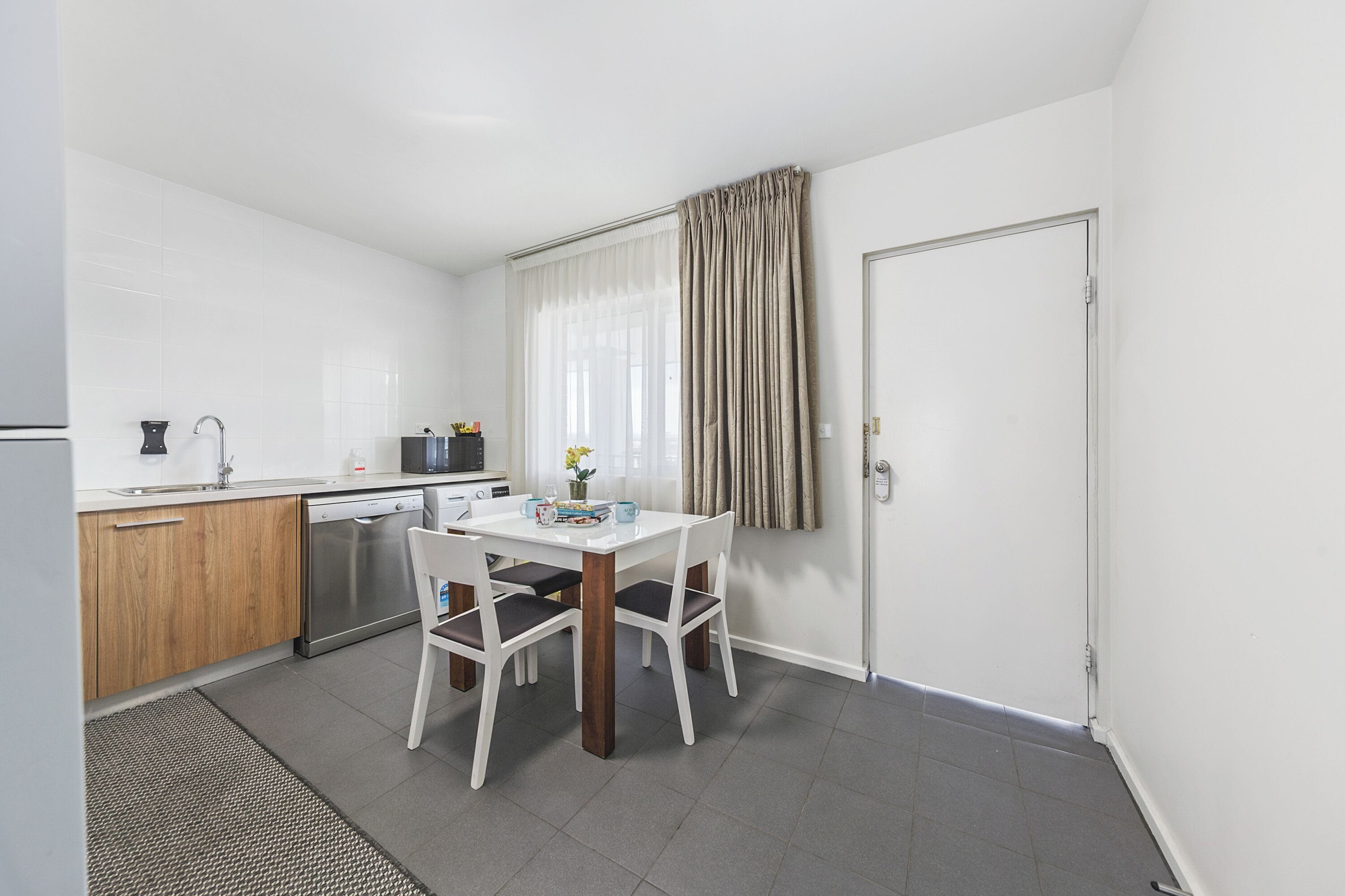 A compact kitchen with a dishwasher, a microwave, and a sink. There is a dining table with chairs and a small window with curtains.