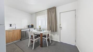 A compact kitchen with a dishwasher, a microwave, and a sink. There is a dining table with chairs and a small window with curtains.