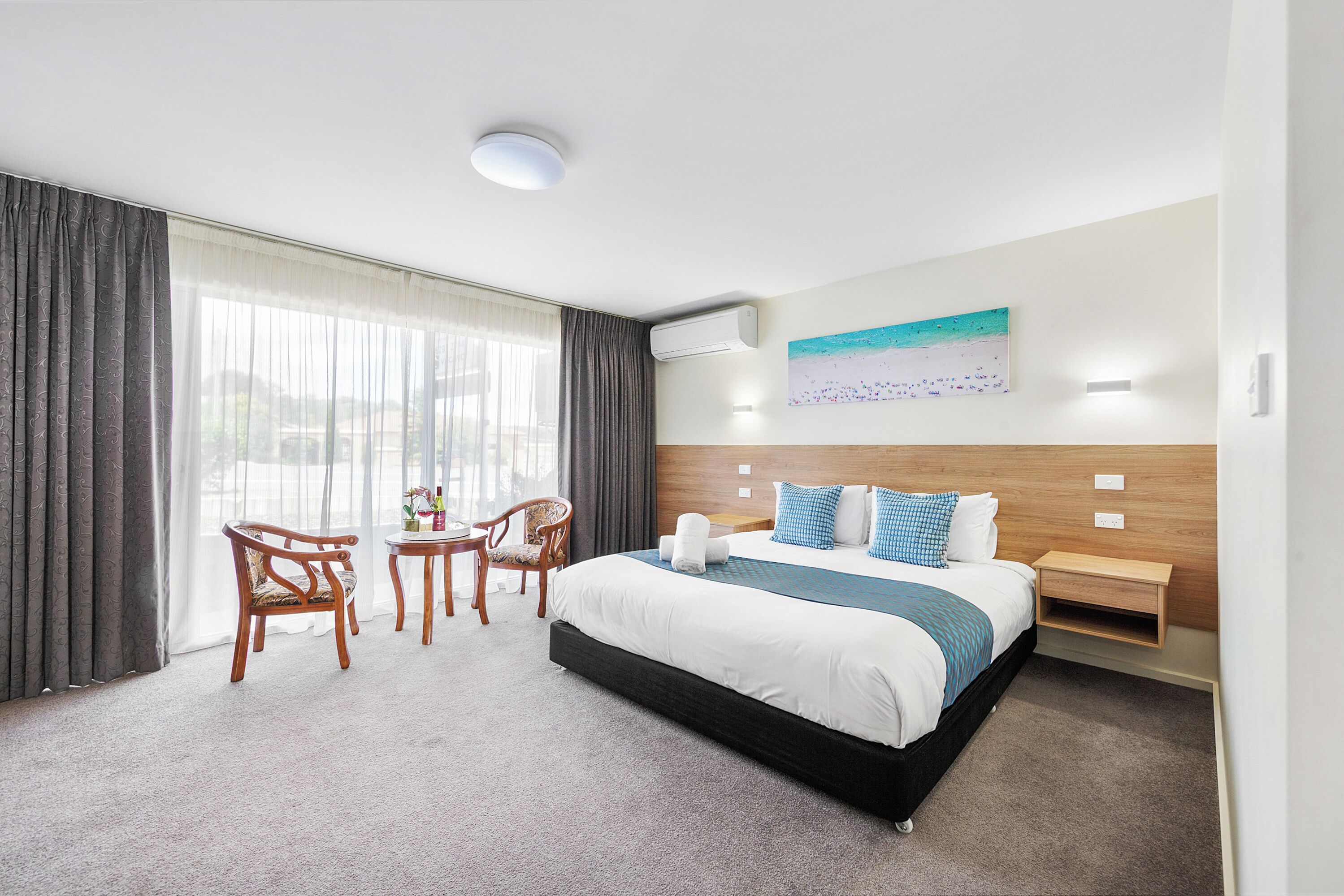 Habitación de hotel con cama, escritorio, dos sillas, ventana con cortinas, unidad de aire acondicionado y un cuadro en la pared.