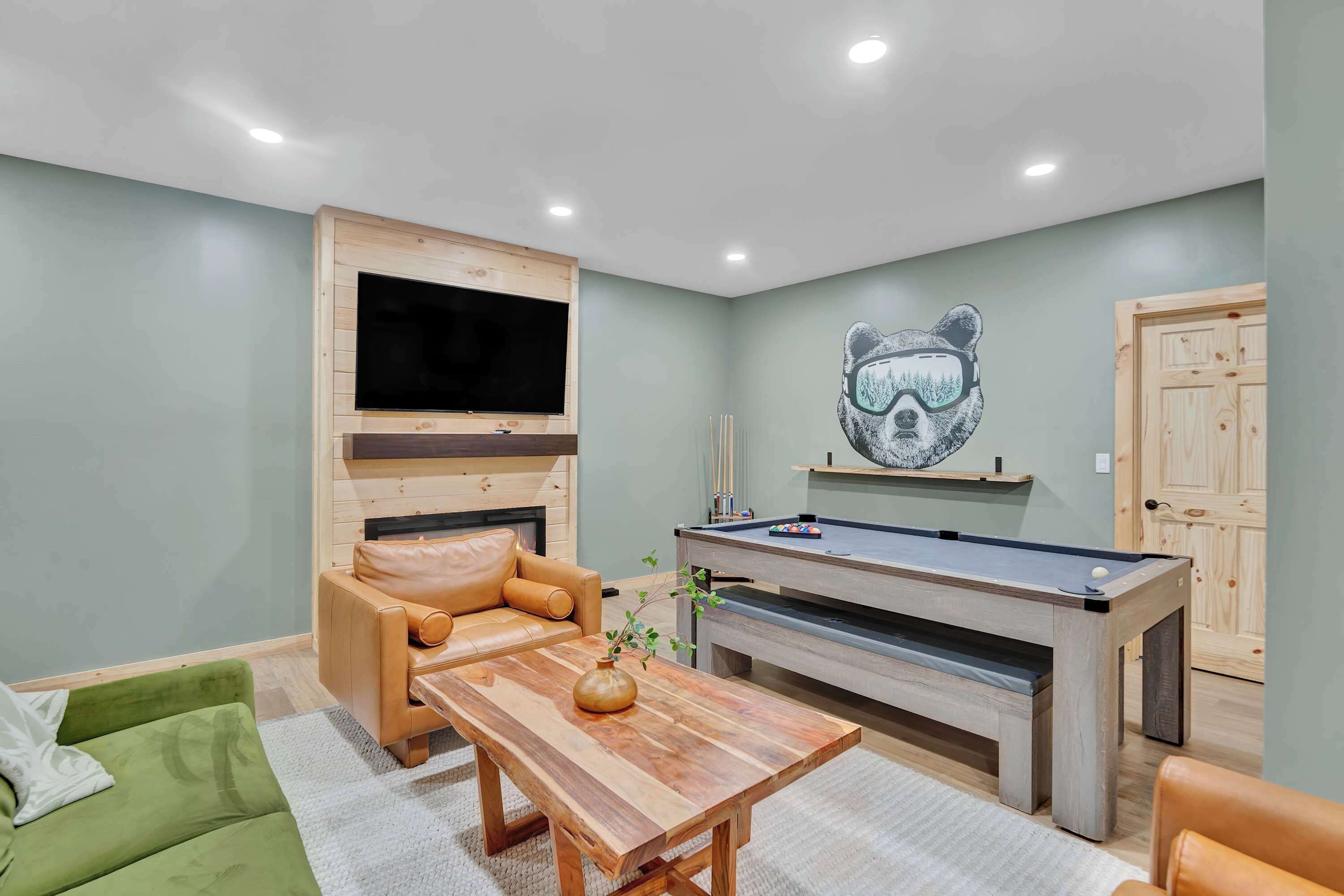 A room with a pool table, a sofa, a TV mounted on a wooden wall, and a bear-themed artwork.