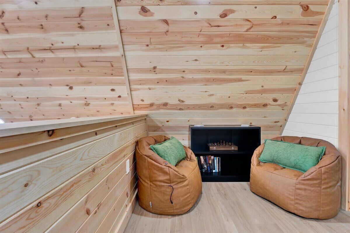 Un acogedor cuarto ático con listones de madera en el techo, dos sillones tipo puff y una estantería negra.