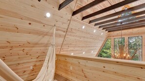 L’intérieur d’un chalet en bois, avec un hamac, des poutres apparentes et une vue sur la forêt par la fenêtre.