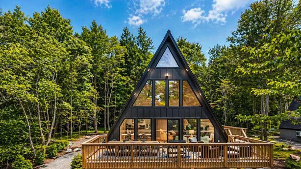 Une cabane moderne en bois, de forme triangulaire, dotée d’une terrasse et entourée d’une forêt dense.