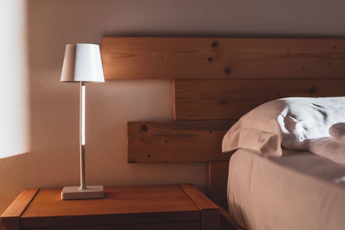A wooden bedside table with a white lamp, a wooden headboard, and a bed with a white pillow.