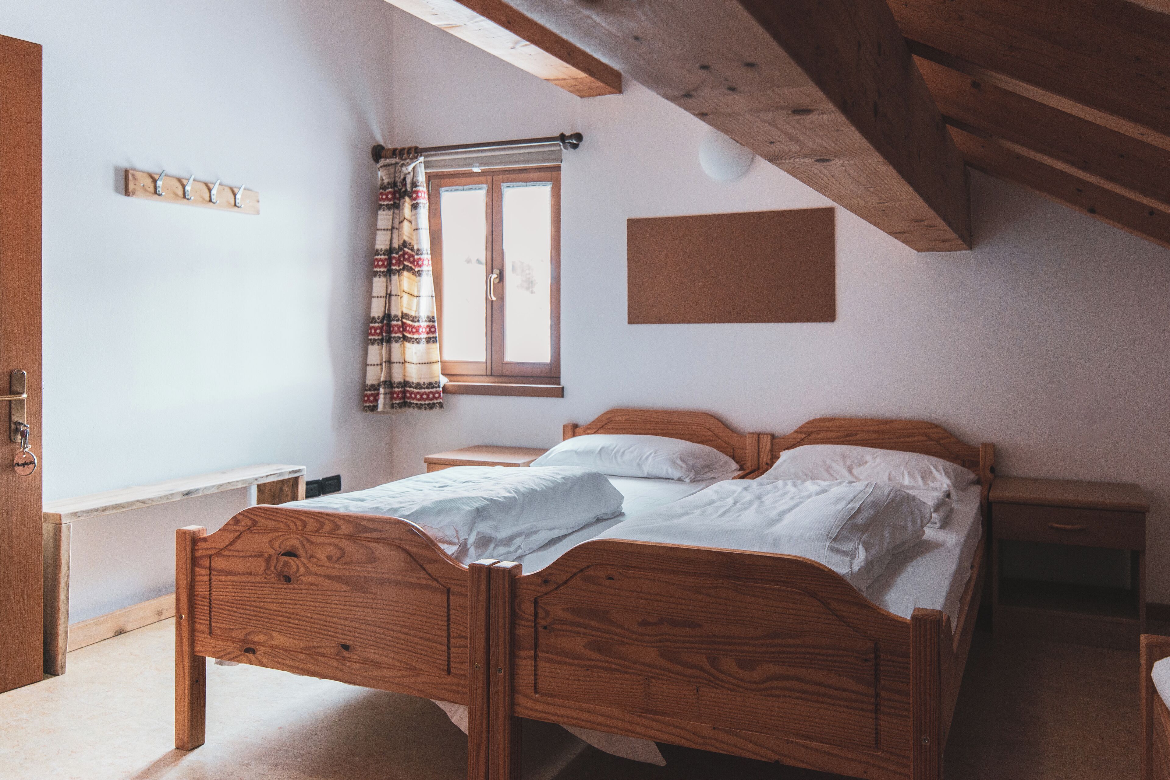 A bedroom with a wooden bed, a window with curtains, a bedside table, and a corkboard on the wall.