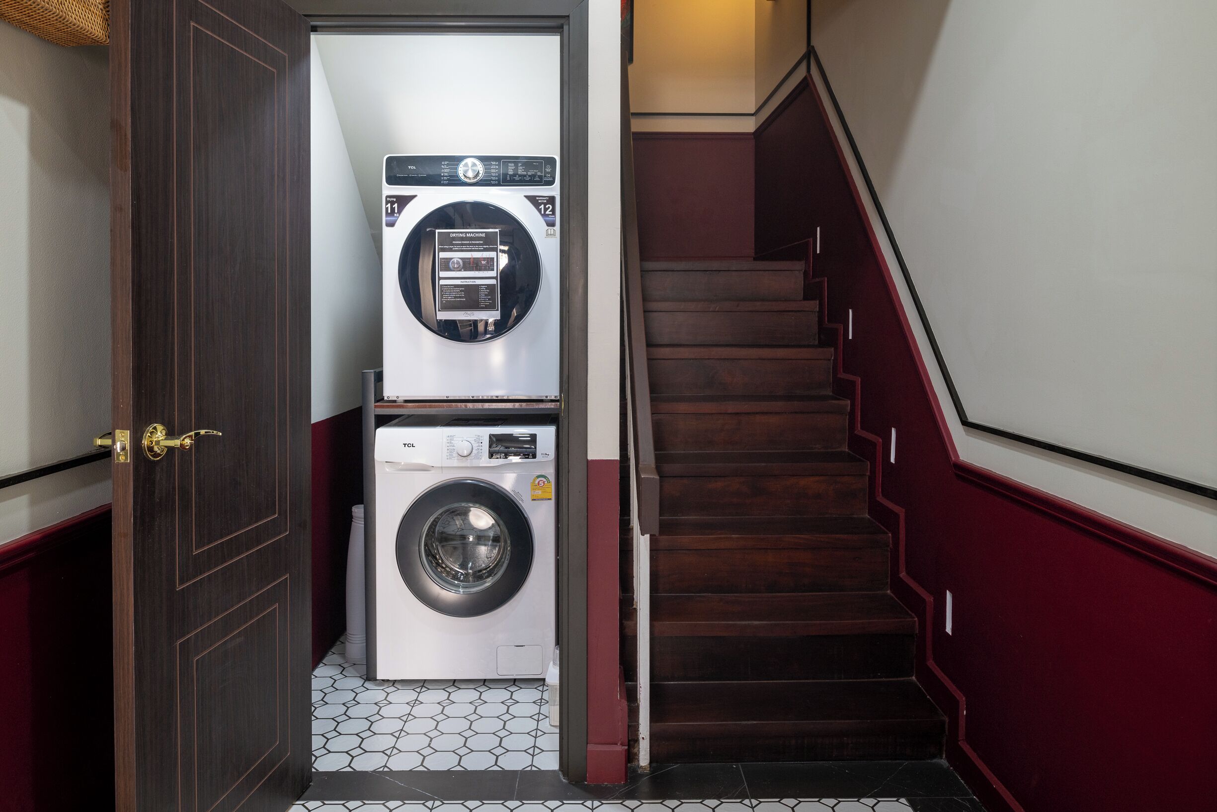A laundry room with two stacked washing machines, a wooden door, and a staircase with red railings.