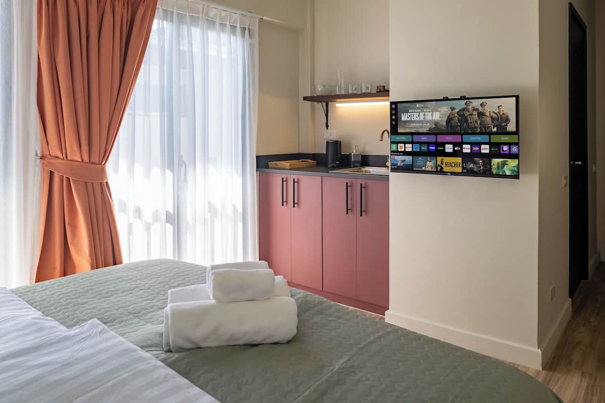 Ein Hotelzimmer mit Bett, einem an der Wand montierten Fernseher mit Filmangeboten, einer Küchenzeile mit Schränken und einem Fenster mit Vorhängen.