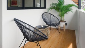 Een balkon met houten vloer, twee zwarte frame stoelen, een plant in pot en een raam met zonwering.