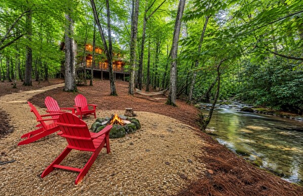 A cabin with a fire pit and red chairs by a stream in a forest.