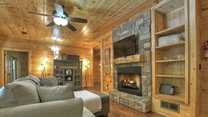 L’intérieur chaleureux d’un chalet avec une cheminée en pierre, des étagères en bois et un ventilateur de plafond.