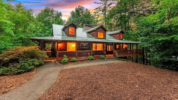 Un chalet rustique doté d’un porche abrité, d’une cheminée en pierre et offrant une vue sur la forêt environnante.