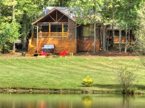 A log cabin with a deck and outdoor seating, surrounded by a grassy area and a body of water. - Pet-friendy, lake-front cabin rental with outdoor fireplace, dock w/Jon Boat (Ellijay)