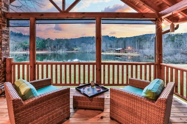 A wooden deck with wicker furniture and a view of a lake and forest. - Pet-friendy, lake-front cabin rental with outdoor fireplace, dock w/Jon Boat (Ellijay)